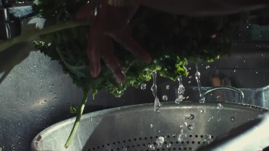 A person's hands carefully wash fresh green kale in a stainless steel sink as water splashes in slow motion