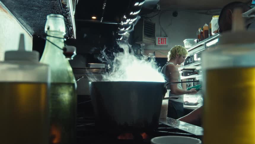Steam billows in slow motion from a large pot on a stove in a dimly lit, professional restaurant kitchen