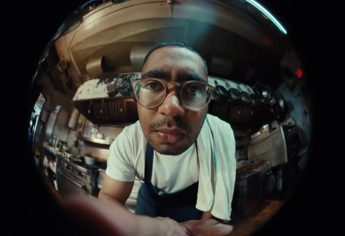 Fisheye POV of a chef in a commercial kitchen who looks into the camera before confidently brandishing a large knife