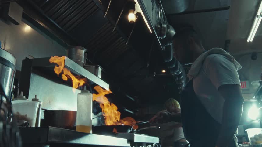 A professional chef cooks with fire in a pan in a dimly lit restaurant kitchen in dramatic slow motion