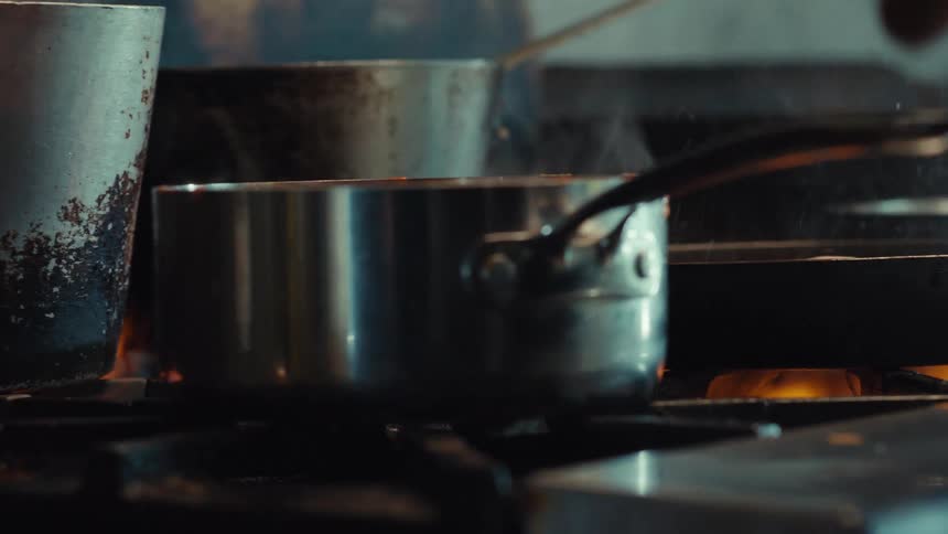 Close up of a chef stirring a simmering sauce in a steel pot over an open flame in a professional kitchen