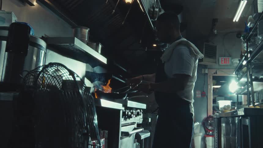 A chef in slow motion skillfully cooks with fire in a dimly lit professional restaurant kitchen