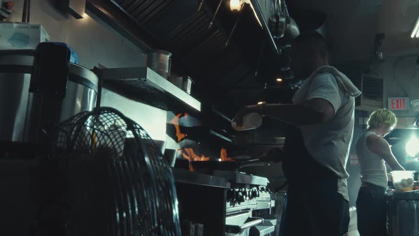 In dramatic slow motion, a chef adds oil to a flaming pan while working in a hot, industrial kitchen