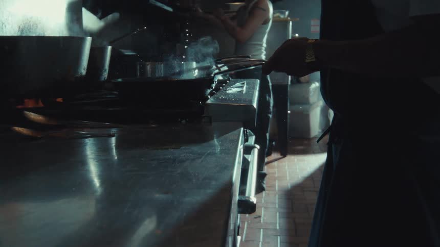 A chef pours oil into a hot pan in a dimly lit restaurant kitchen, creating a dramatic sizzle in super slow motion