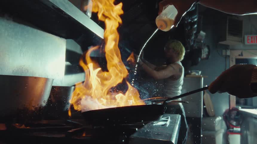 A chef pours liquid into a hot pan, causing a dramatic fire flare-up in super slow motion