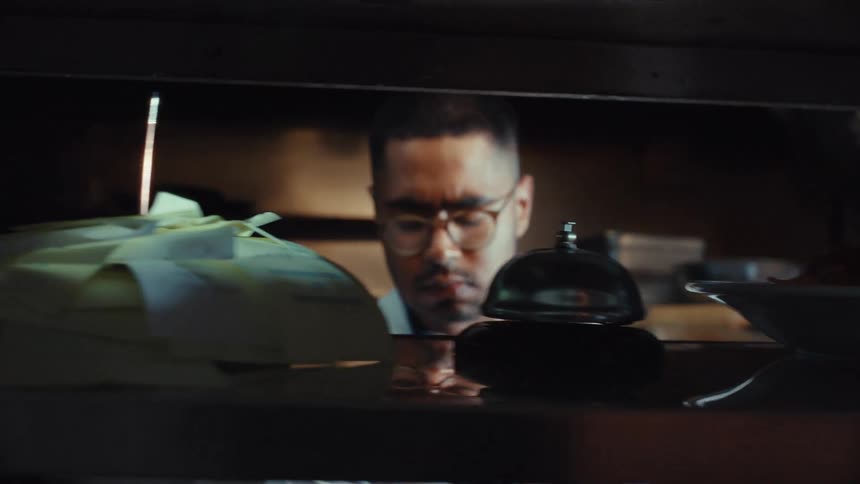 Tracking shot of a restaurant's food service window where hot dishes are lined up and orders are taken by chefs in a dimly lit kitchen
