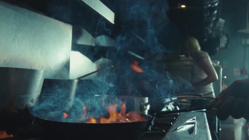A dramatic slow-motion close-up of a chef flambéing food in a pan on a restaurant stovetop