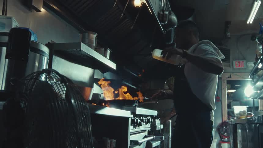 A skilled chef expertly flambés a dish in a professional kitchen, with flames erupting from the pan in dramatic slow motion