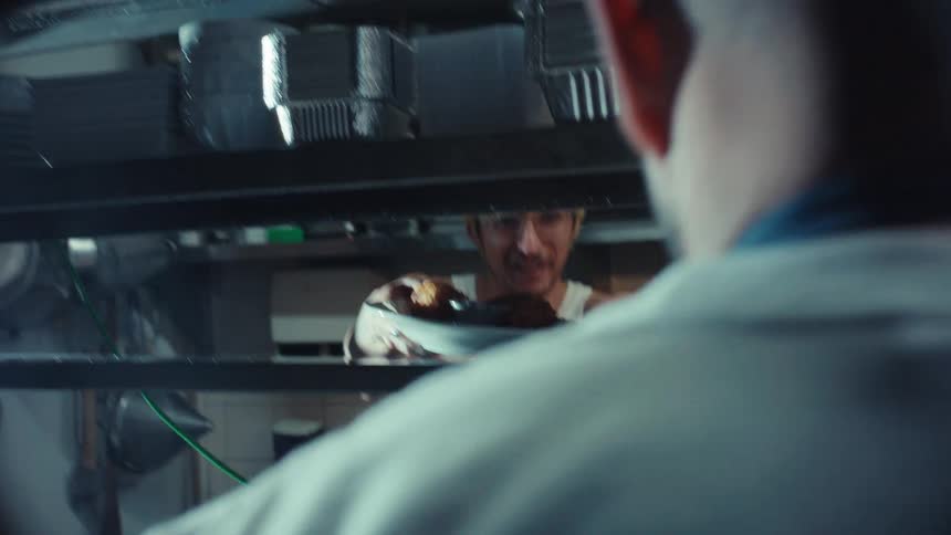 Over the shoulder shot of a chef passing a plated dish to a waiter in a professional restaurant kitchen