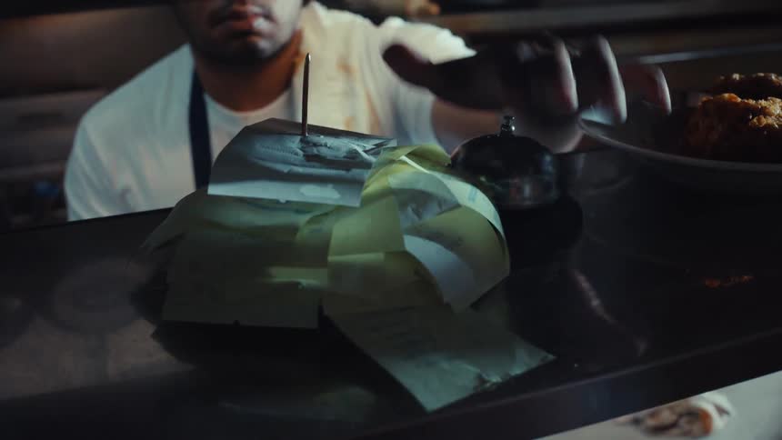 A close-up of a pile of order tickets grows as a busy chef rings the service bell in the background of a restaurant kitchen
