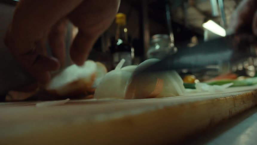 A dramatic low-angle close-up shot of a chef's hands skillfully slicing a white onion on a cutting board in a kitchen