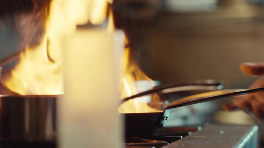 A professional chef expertly flambés food in a hot pan, causing a dramatic burst of flames in a commercial kitchen