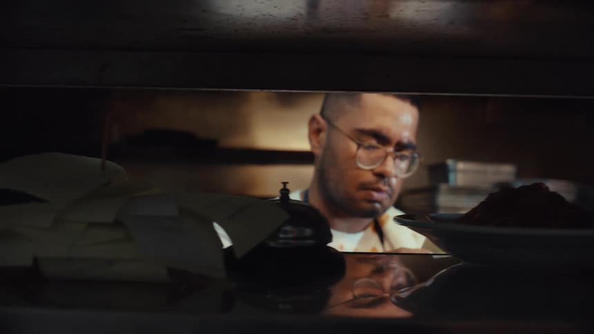 A busy chef rings the service bell and puts an order ticket on a spike in a dimly lit commercial kitchen