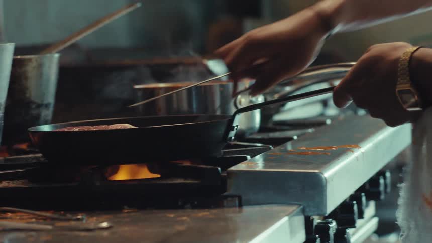 Close-up slow-motion shot of a focused chef cooking a steak in a sizzling frying pan over an open flame in a professional kitchen