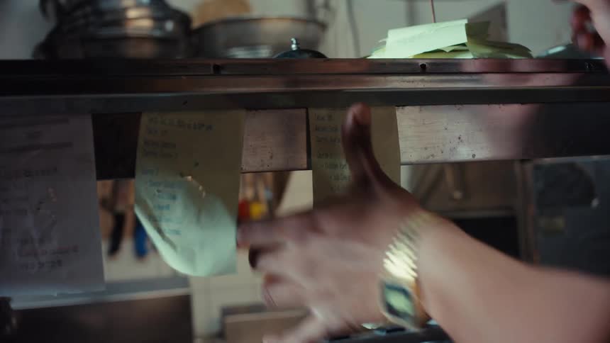 A cook's hands place a new order ticket on the rail in a commercial restaurant kitchen