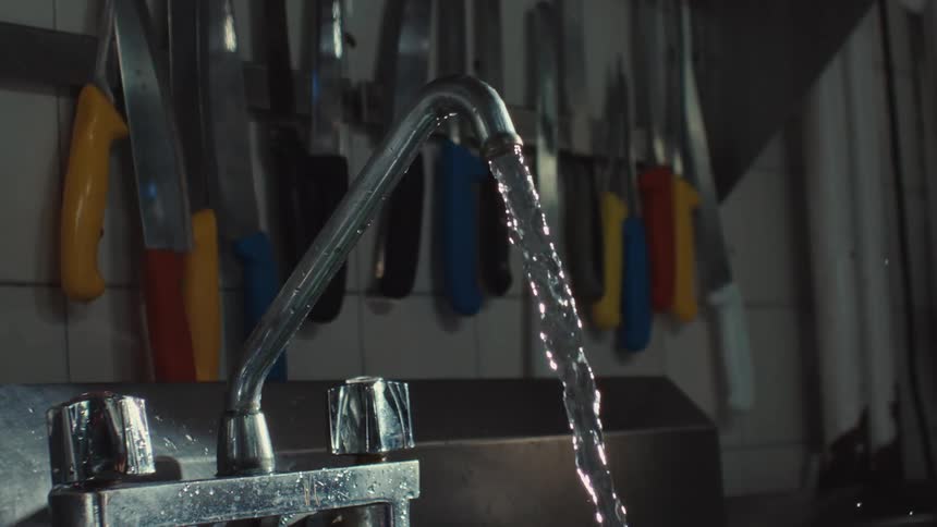 Water pours in slow motion from a stainless steel faucet in a commercial kitchen with knives on the wall