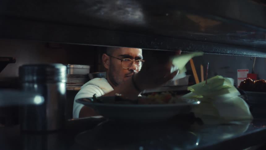 Exhausted chef wipes sweat from his face while preparing an order in a dimly lit, hot restaurant kitchen