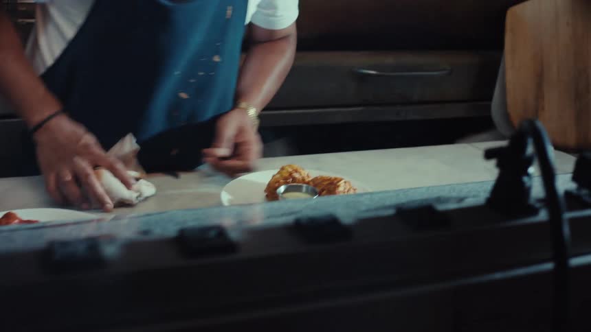 A focused chef meticulously plates food in a dimly lit, bustling professional restaurant kitchen as seen through the service window