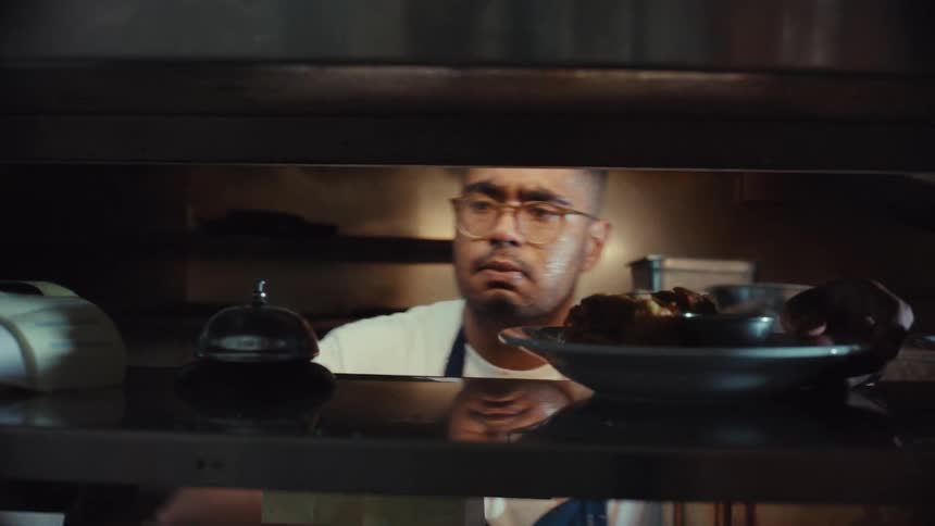 A focused chef prepares multiple dishes, places them on the serving pass, and rings the bell in a dimly lit, busy restaurant kitchen