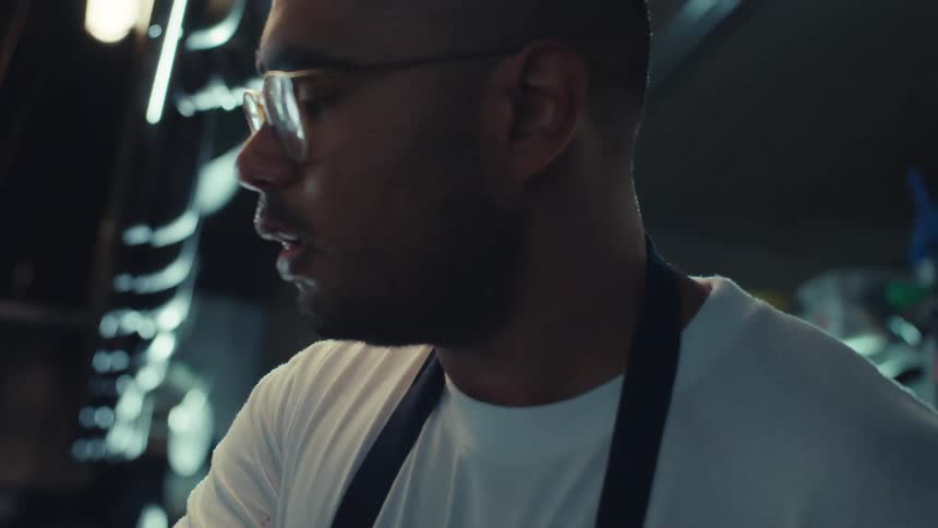 A focused chef in an apron tastes his food and sautés ingredients in a pan on a gas stove in a bustling restaurant kitchen