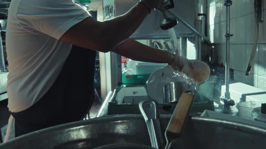A restaurant worker in an apron rinses a dirty dish in a metal sink using a high-pressure spray nozzle in slow motion
