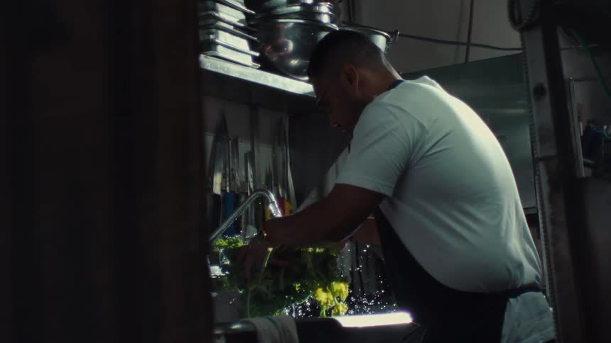 A cook in a dimly lit professional kitchen carefully washes fresh green vegetables in a sink in slow motion