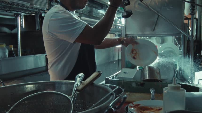 Slow motion shot captures a dishwasher using a high-pressure sprayer to clean a dirty plate in a restaurant kitchen