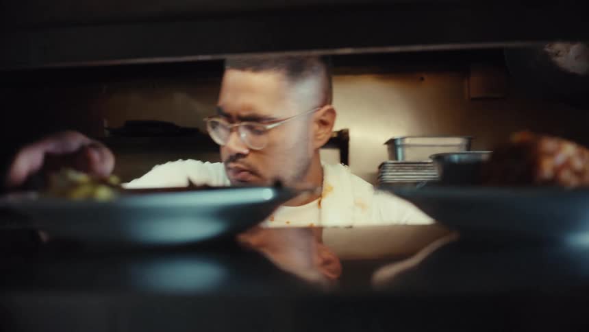 Slow motion close up of a professional chef plating delicious meals and wiping the plate edges in a busy commercial kitchen