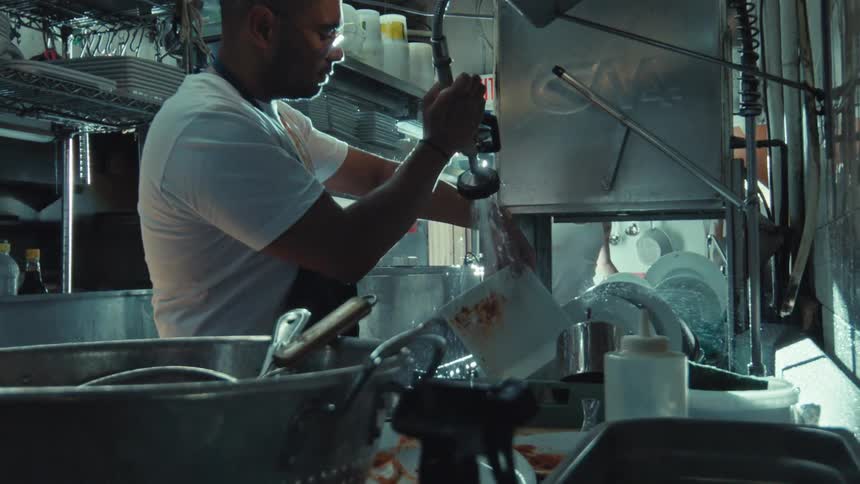 A dishwasher in a dimly lit commercial kitchen meticulously rinses a dirty plate in slow motion
