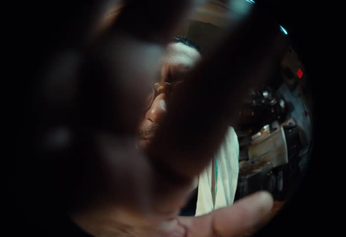 A chef in a commercial kitchen looks closely into a fisheye lens, then playfully brandishes a large knife at the camera