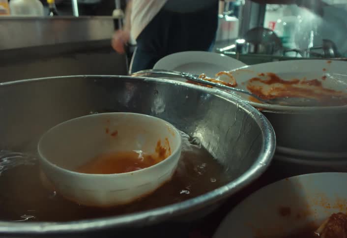 A dirty bowl is thrown into a basin of water in slow motion in a busy restaurant kitchen