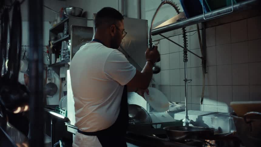 A restaurant employee works at the dishwashing station, rinsing dirty plates before loading them into a commercial dishwasher in a busy kitchen
