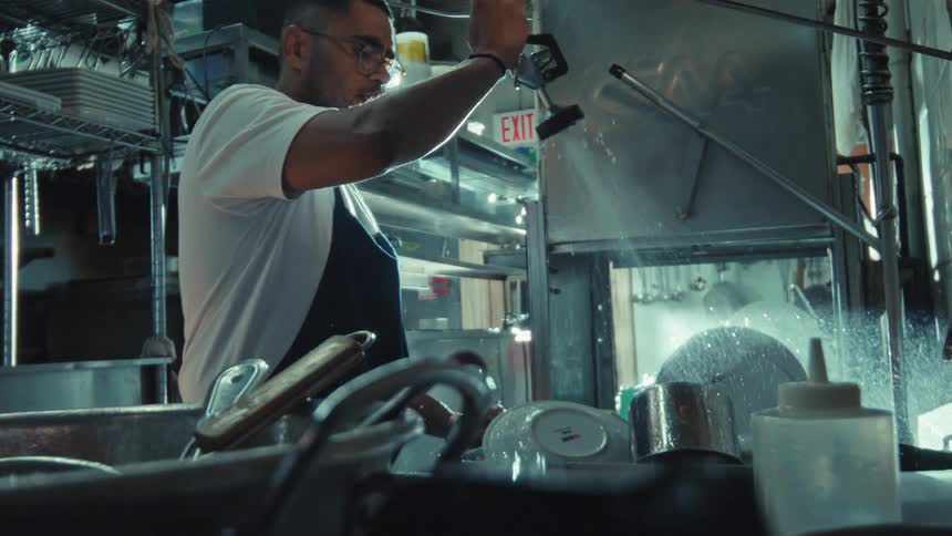 A kitchen worker sprays down dishes with a high-pressure nozzle in a commercial restaurant sink in cinematic slow motion