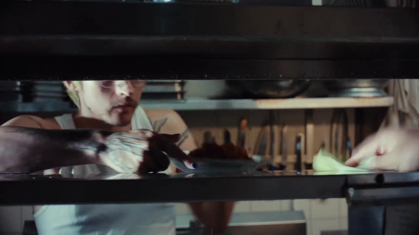 A bustling restaurant kitchen seen from the pass as one chef cleans and another hands off freshly plated dishes