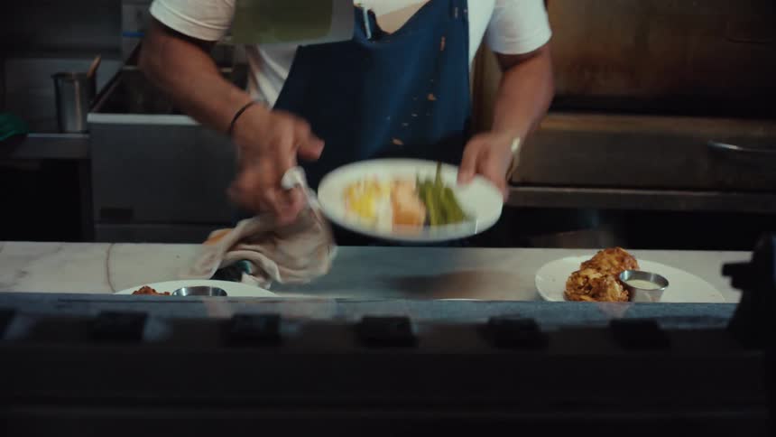 A focused chef meticulously prepares and plates multiple dishes, wiping them clean before ringing the service bell for pickup in a restaurant kitchen