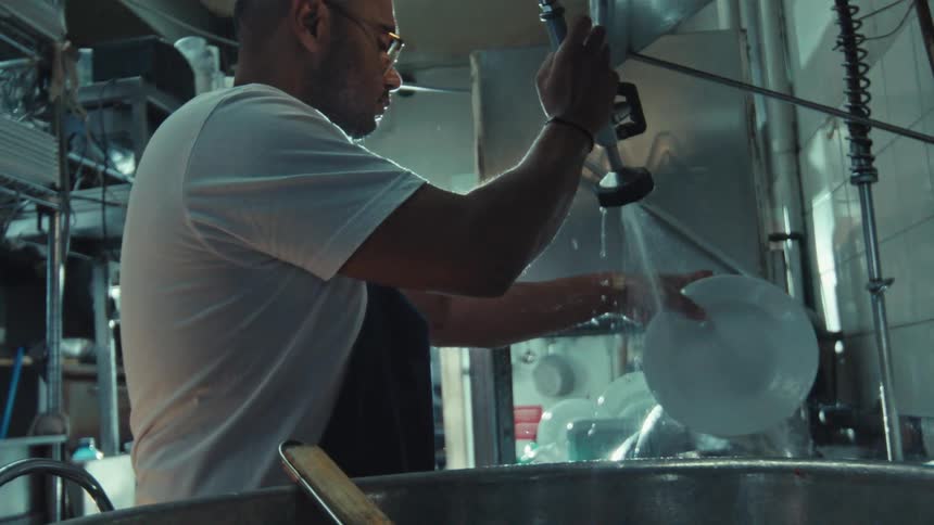 A male restaurant worker in an apron rinses a white plate with a high-pressure water sprayer in a commercial kitchen, filmed in slow motion