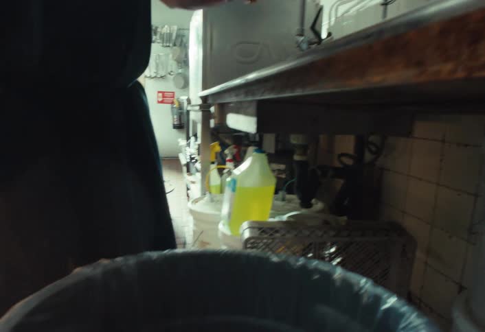 A kitchen worker scrapes leftover spaghetti and meatballs off a plate and into a garbage can in a restaurant kitchen