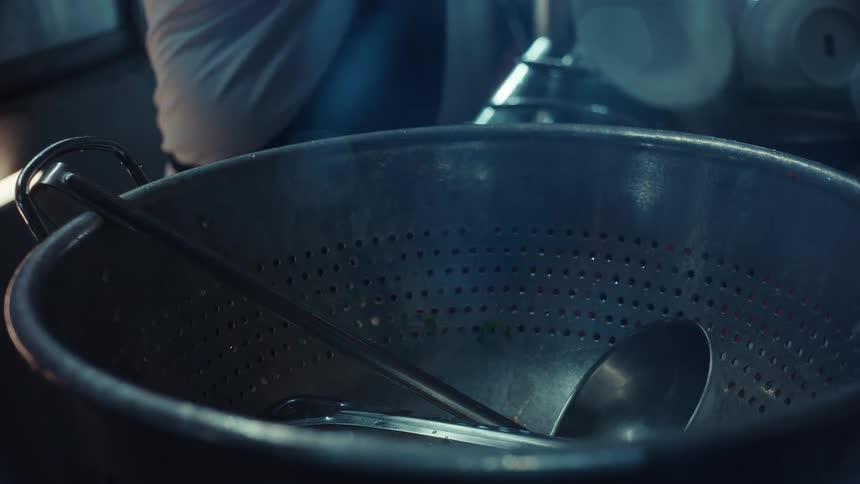 A kitchen worker removes washed bowls and utensils from a large metal colander in a dimly lit setting