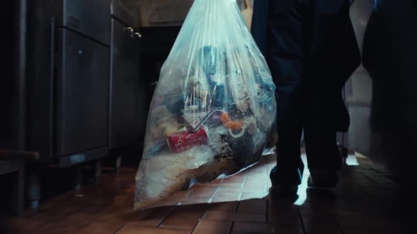 A kitchen worker carries a large, full bag of trash through a restaurant kitchen floor in slow motion