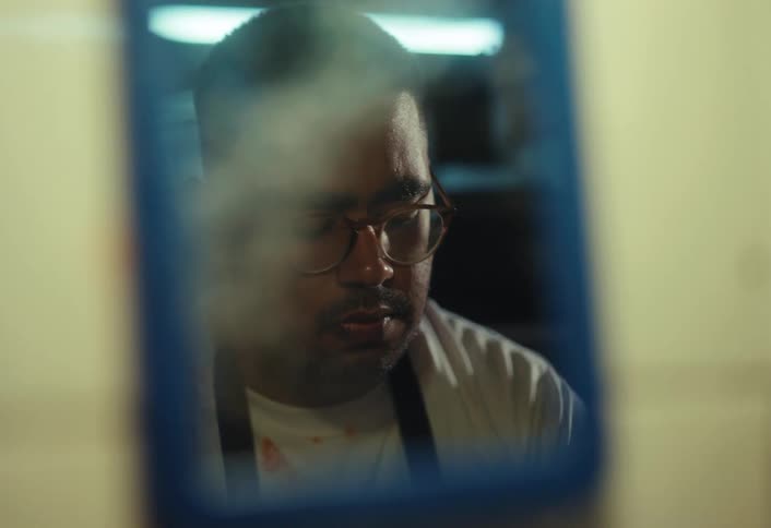A tired man with glasses contemplates his reflection in a small, steamy mirror, then slowly closes his eyes
