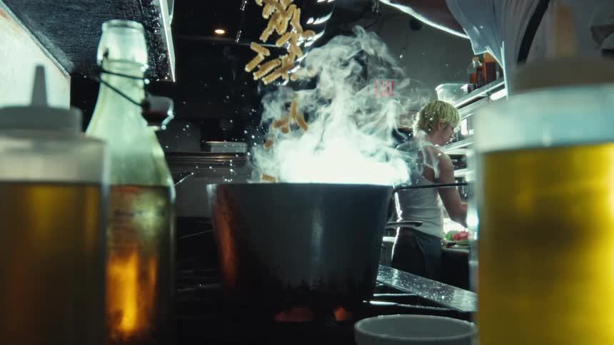 A chef tosses pasta into a steaming pot of boiling water in a bustling restaurant kitchen in dramatic slow motion