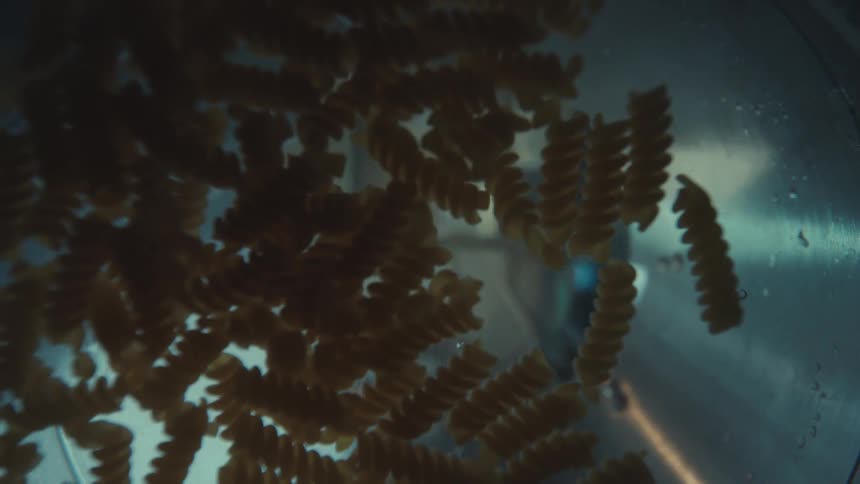 A man pours fusilli pasta into a pot of boiling water, captured from a unique perspective inside the pot