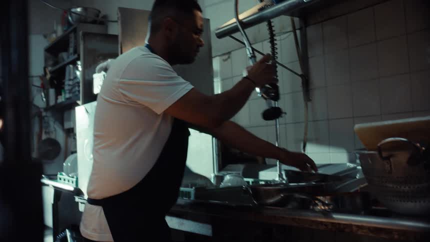 A diligent male dishwasher meticulously cleans pots and pans with a high-pressure spray hose in a dimly lit industrial kitchen