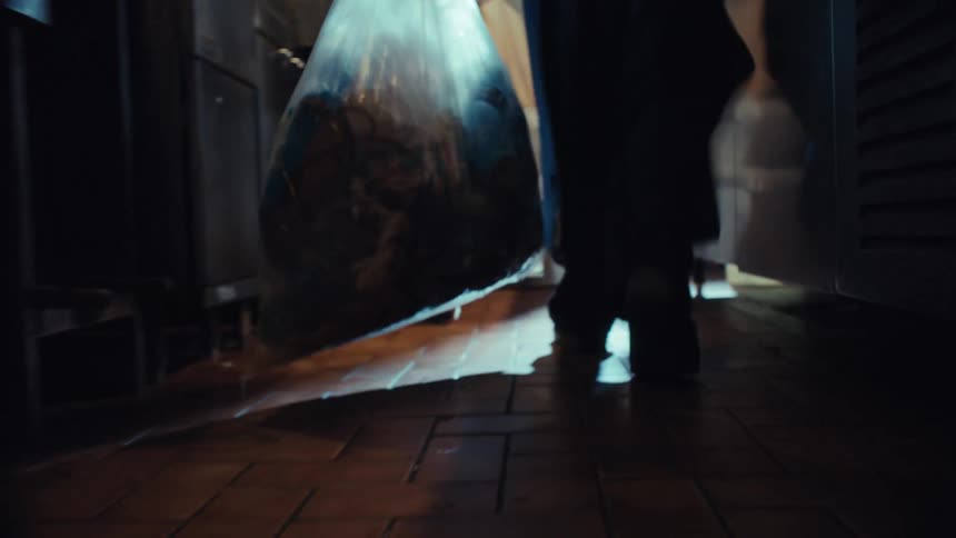 A low-angle slow-motion shot follows a person dragging a leaking trash bag across a dark, wet kitchen floor