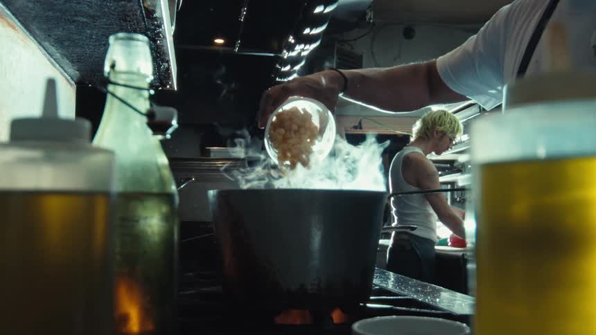 A chef pours pasta into a steaming pot in a dimly lit restaurant kitchen in dramatic slow motion