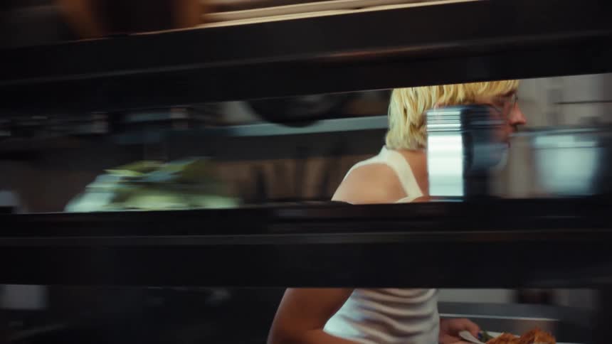 A tattooed chef with blond hair rushes through a dimly lit restaurant kitchen, grabbing finished food plates from the pass