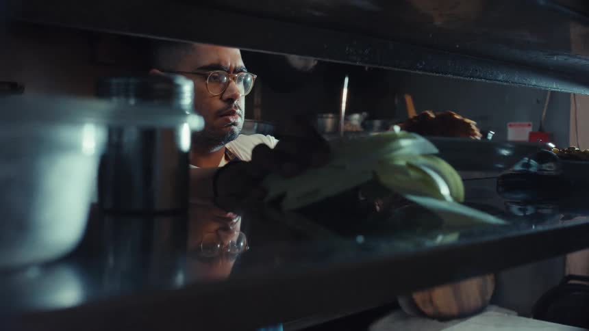 A male chef wearing glasses carefully plates a dish in a dimly lit professional restaurant kitchen
