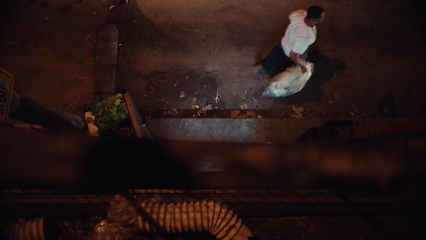 Top-down view of a man dragging a full trash bag down a dirty city street at night