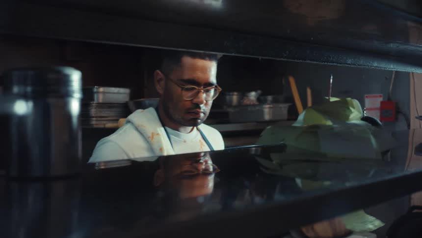 A focused chef wearing glasses grabs an order ticket in a bustling restaurant kitchen at night