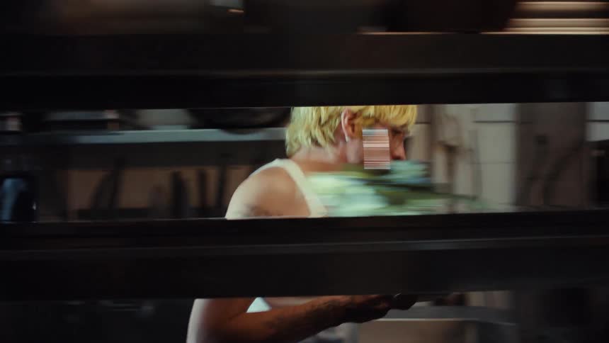 View from the pass of a tattooed chef in a tank top grabbing two plates of food and walking through a dimly lit restaurant kitchen